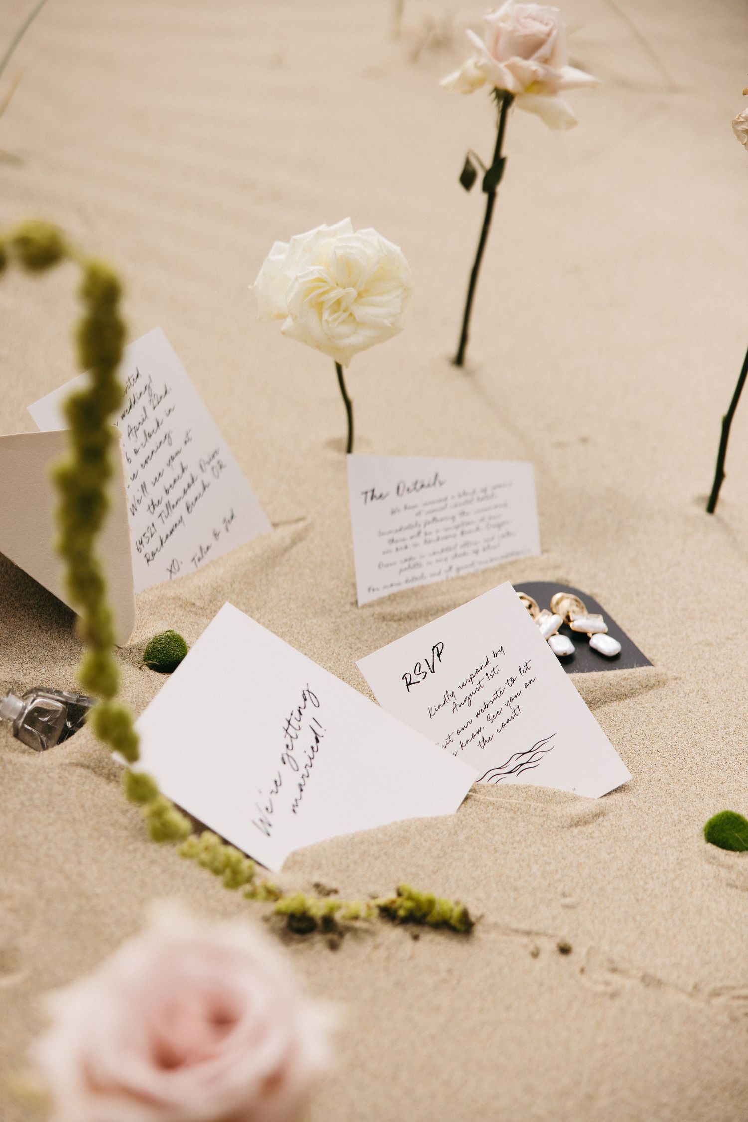 Wedding RSVP cards and white flowers arranged on beige textured surface with green moss accents.