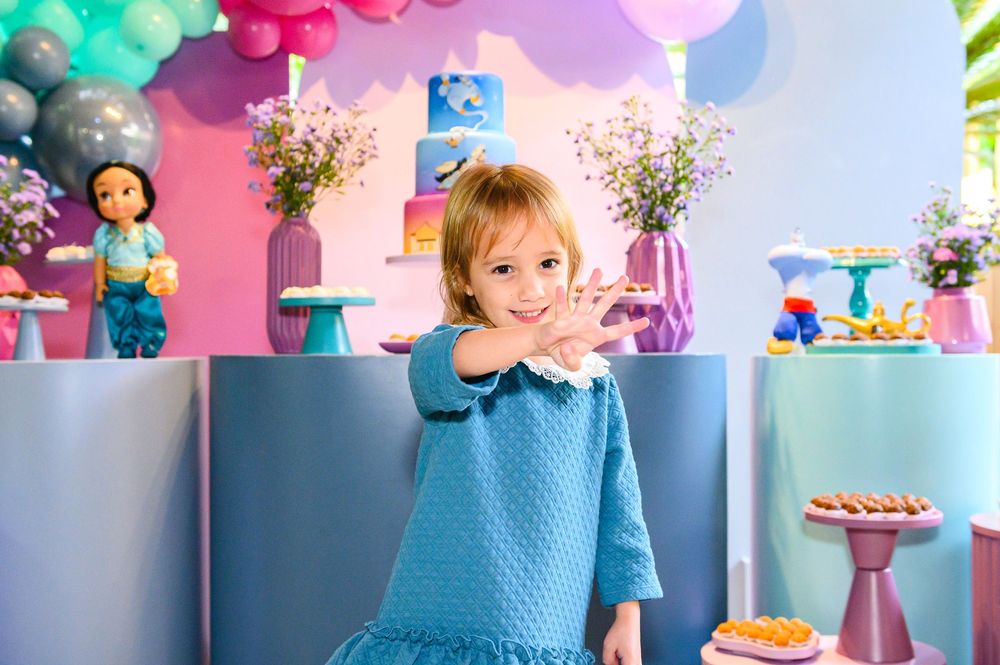 Child in blue dress smiles at birthday party with pink and mint balloon decorations and dessert table in background.
