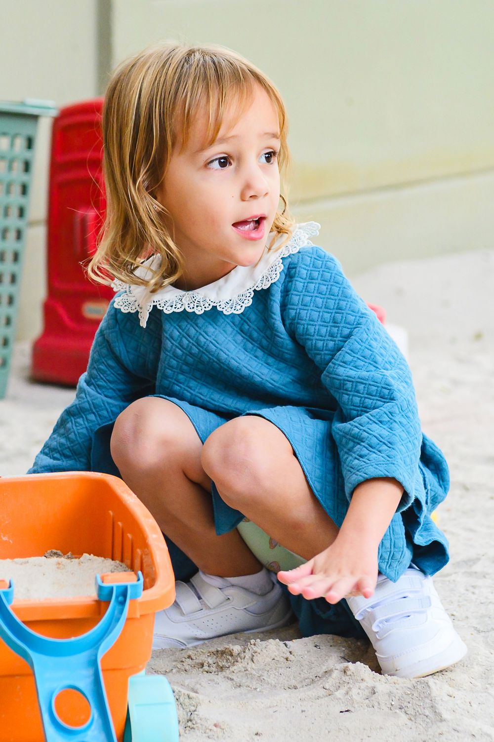 Child wearing blue dress plays with colorful sand toys at a beach or sandbox.