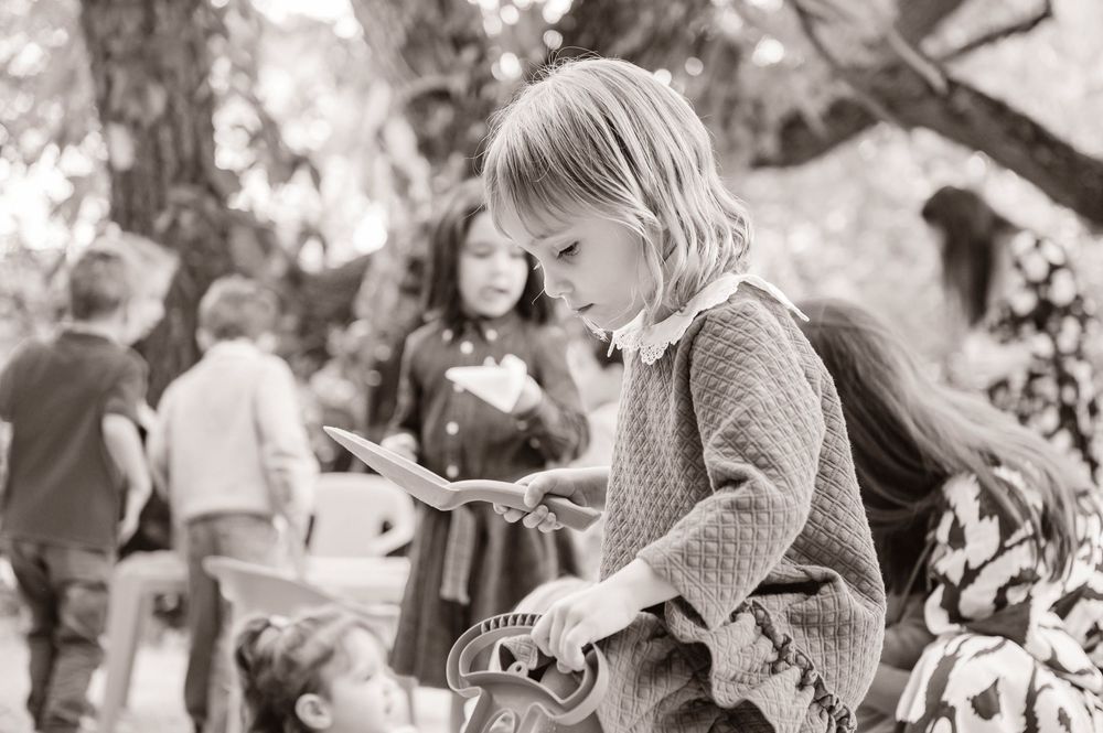 A child in a knit sweater sits outdoors while others gather in the background in this black and white photograph.