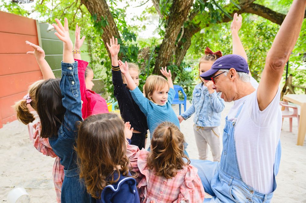 A group celebrates outdoors with raised arms and joyful expressions beneath green tree branches.