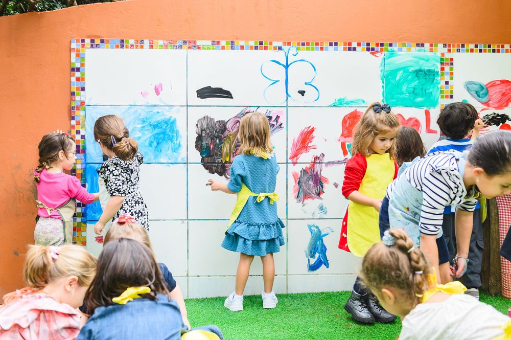 A group of children looking at drawings posted on an orange wall during an educational activity outdoors.