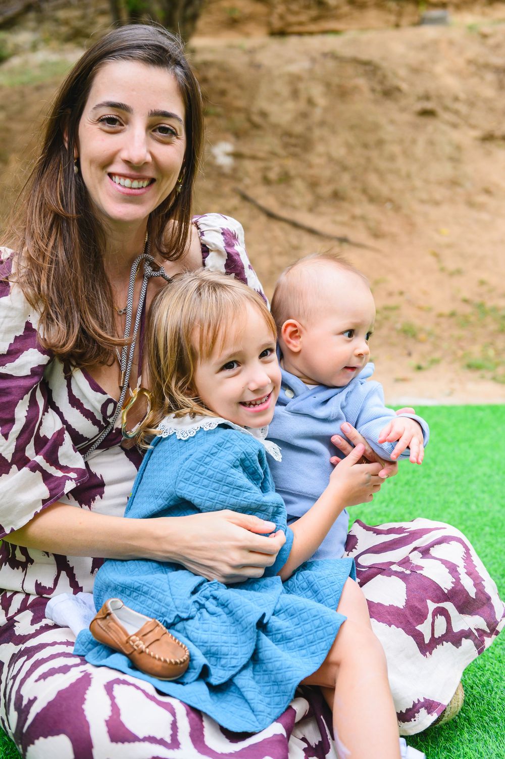 Three family members sit together on grass outdoors, wearing coordinated blue and purple outfits on a sunny day.