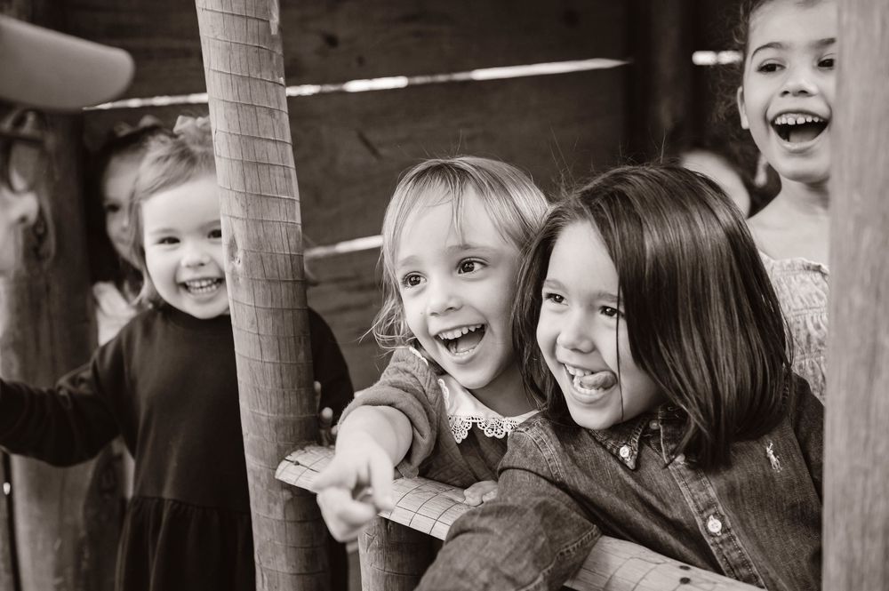 Black and white candid photograph of joyful expressions and laughter captured through wooden fence posts.