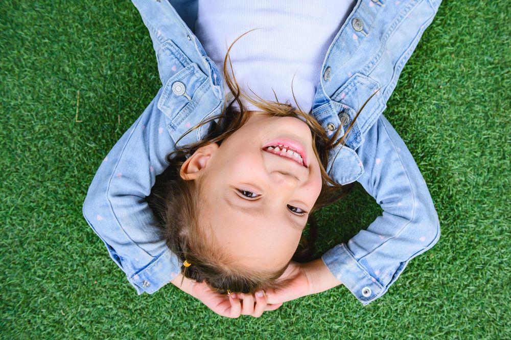 Person lying on green artificial turf wearing light denim clothing and smiling while looking up at the camera.
