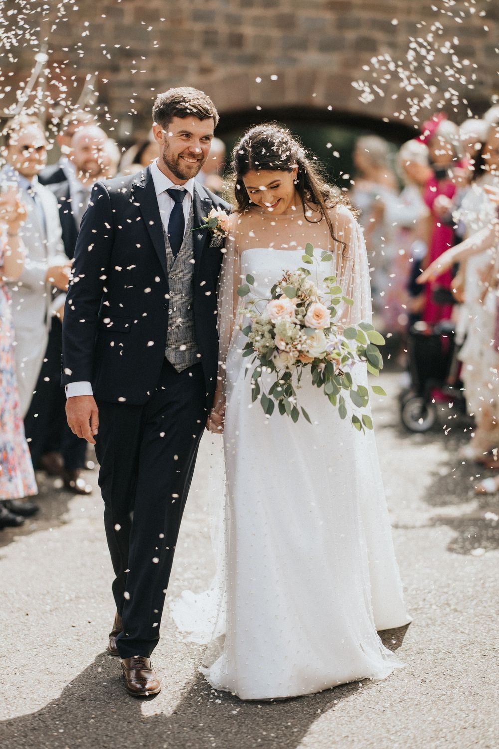 Newlyweds walk together under shower of confetti while wedding guests celebrate in background at outdoor ceremony.