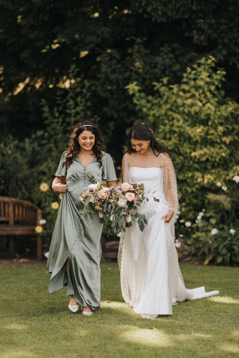 Two women in elegant dresses, one sage green and one white, walk together on a garden lawn holding bouquets.