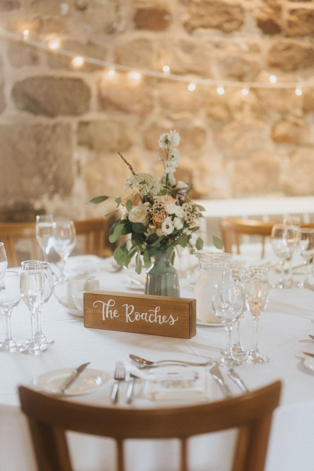 Elegant wedding table setting with wooden name card holder, floral centerpiece and string lights against stone wall background.