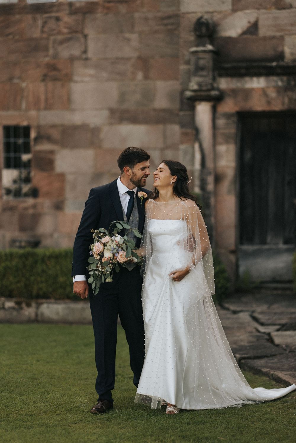 A romantic wedding couple stands on castle grounds with stone walls and lush grass in a moody atmosphere.