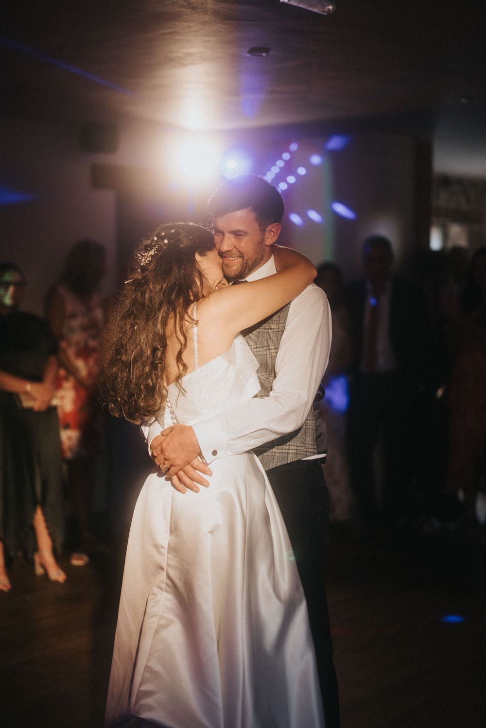 A romantic wedding dance moment captured on a dimly lit dance floor with blue lights and white formal attire.