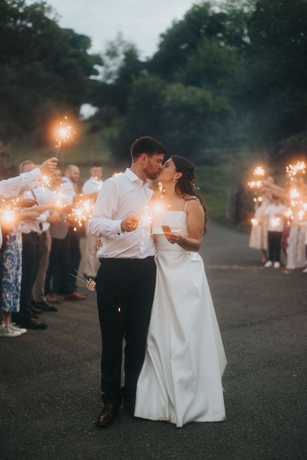Newlyweds share romantic moment with sparklers during evening wedding send-off with guests in background.