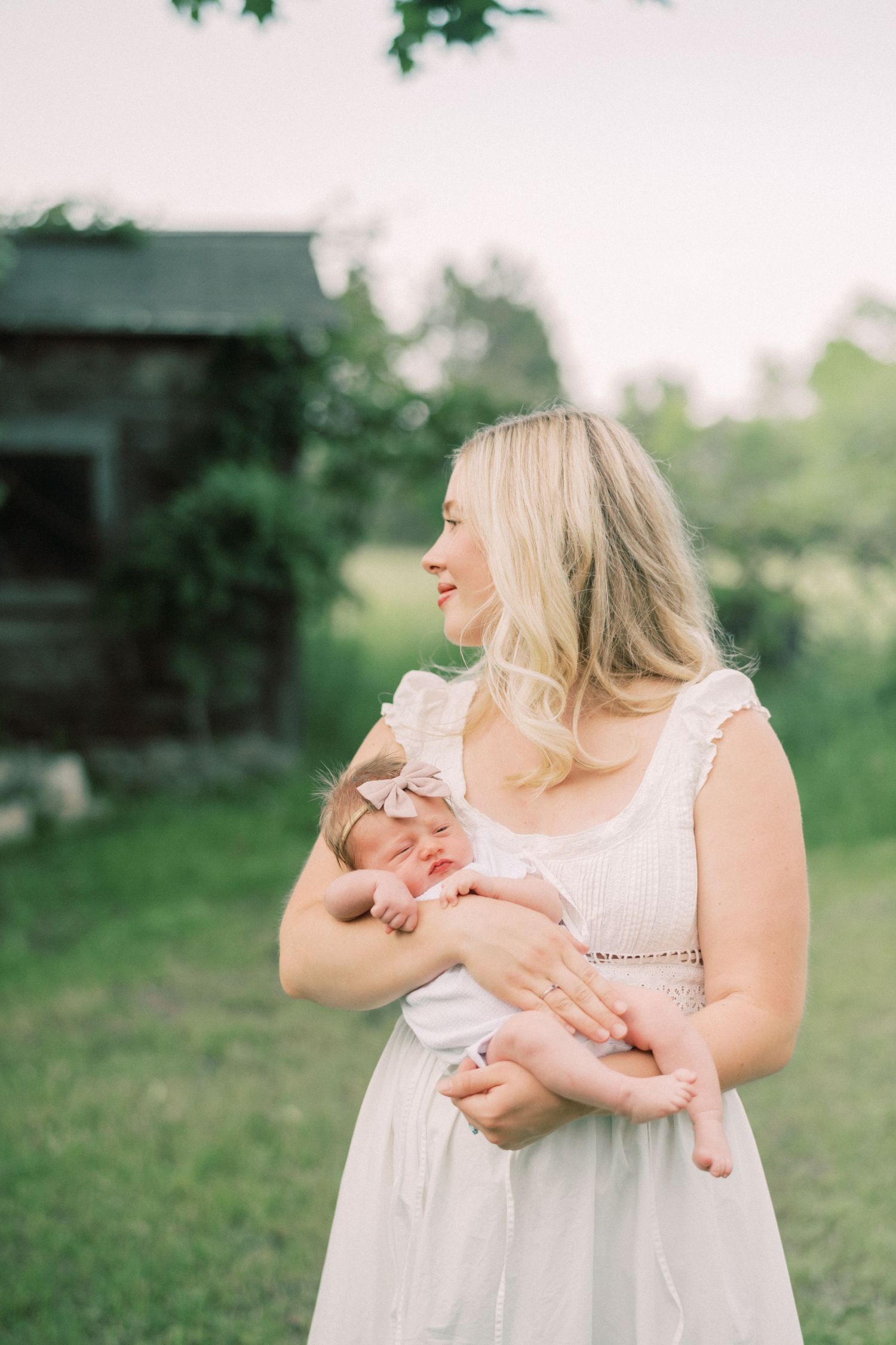 A mother in a white dress holds her newborn baby in a tender outdoor photoshoot at sunset.