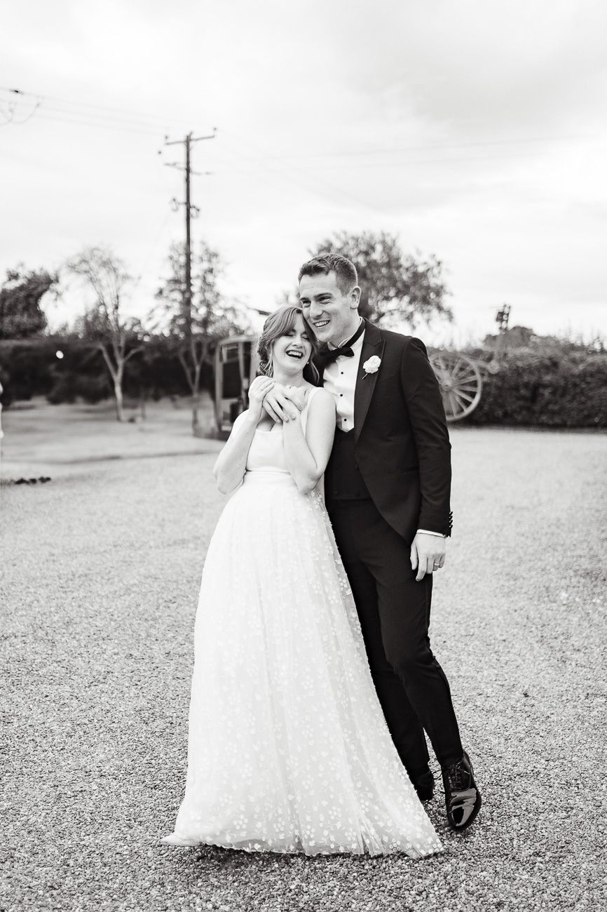A bride and groom in formal wedding attire walk along gravel outdoors in a black and white photo series.
