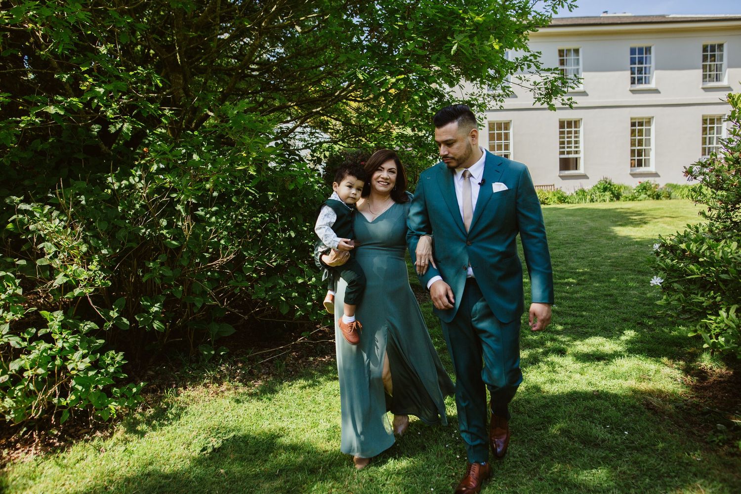 People in teal and turquoise attire walking and laughing together in a lush garden setting near a white building.