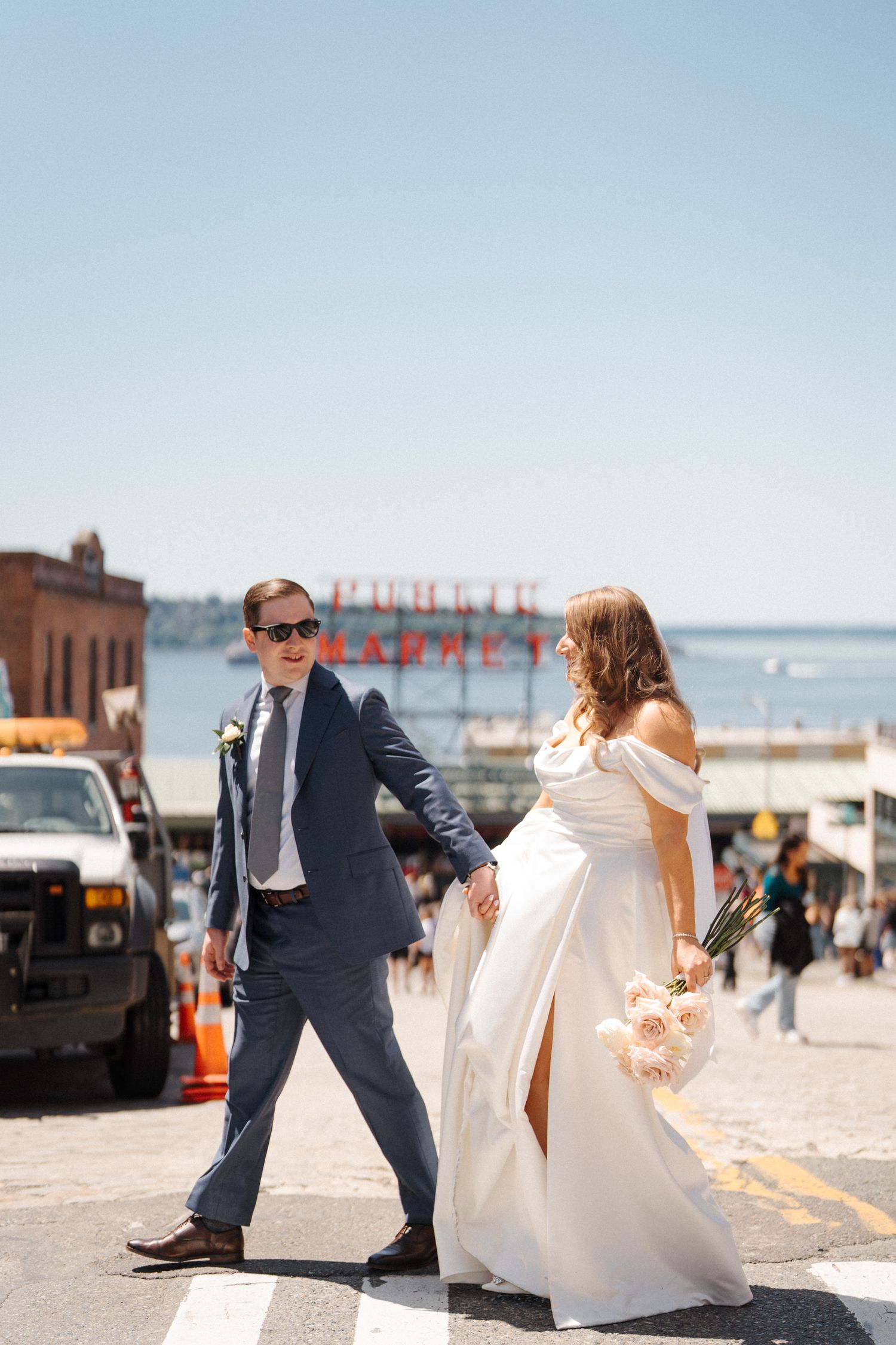 A couple shares a playful dance moment on a sunny beachside boardwalk in formal wedding attire.