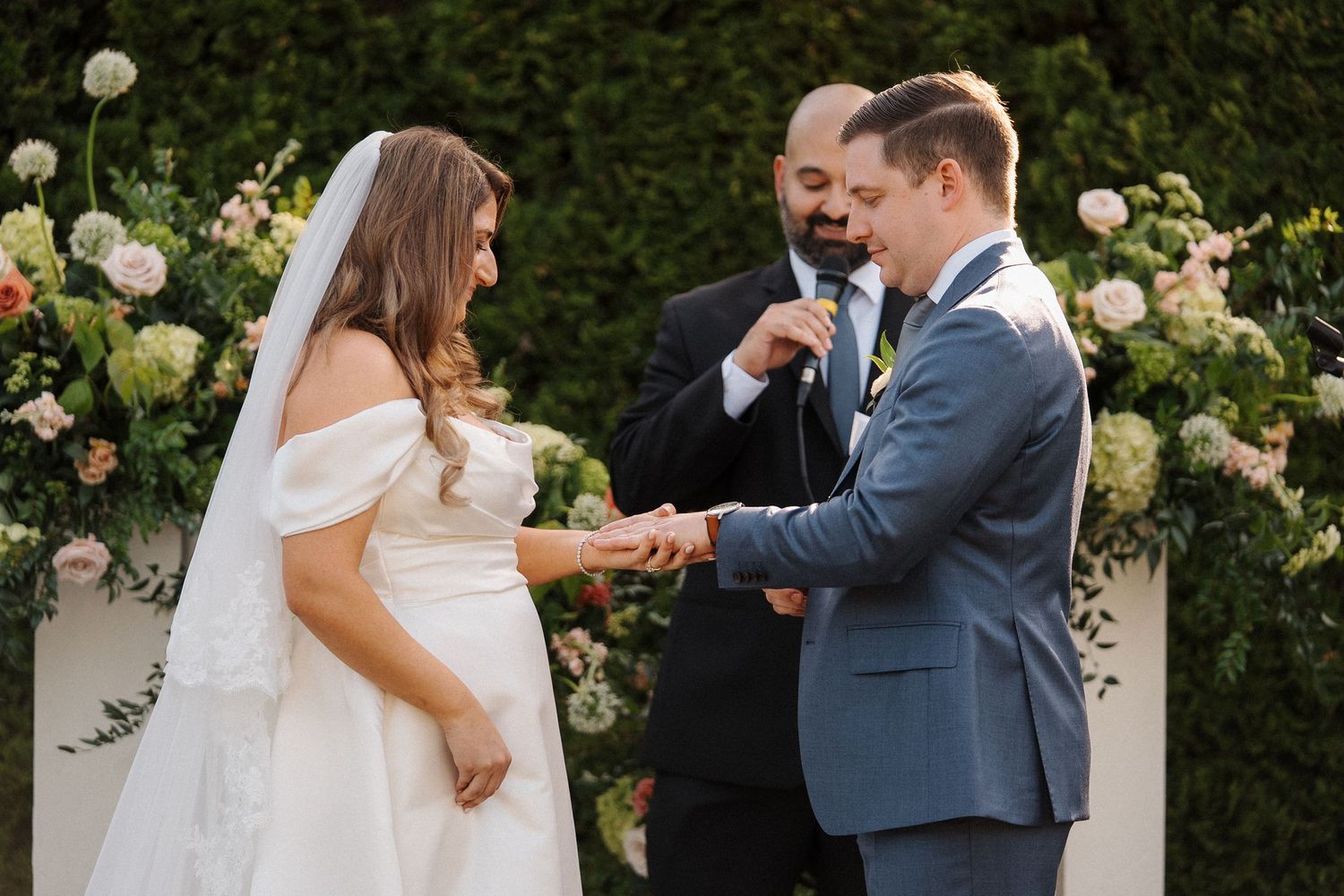 A couple exchanges wedding rings during an outdoor ceremony surrounded by blooming yellow roses and white flowers.