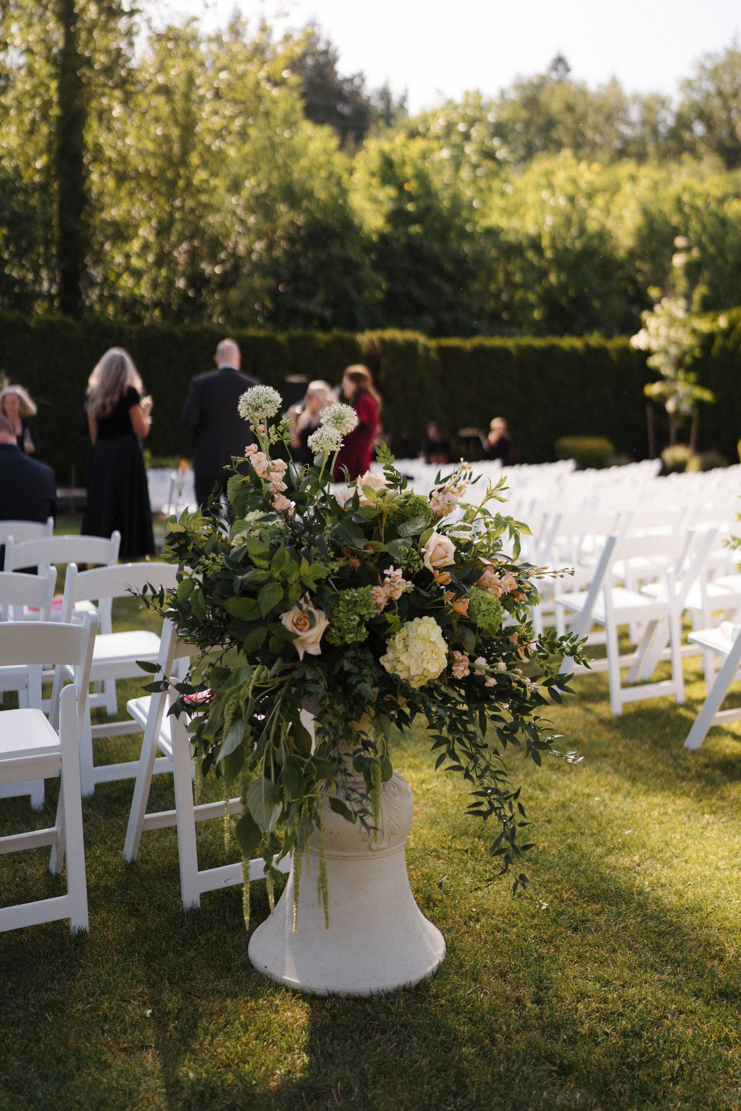 A large floral arrangement in white and pink sits on a pedestal at an outdoor wedding ceremony with white chairs on a lawn.