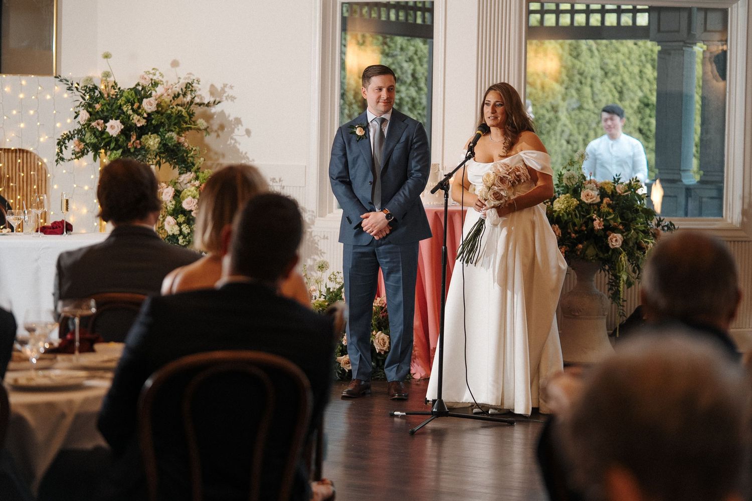 Two people stand at the front of a room decorated with floral arrangements during an indoor celebration.