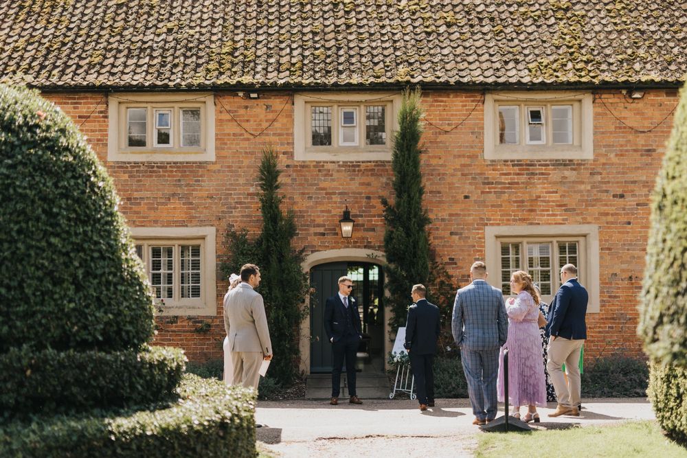 A group of people standing outside a rustic brick country house entrance with tall cypress trees framing the doorway.