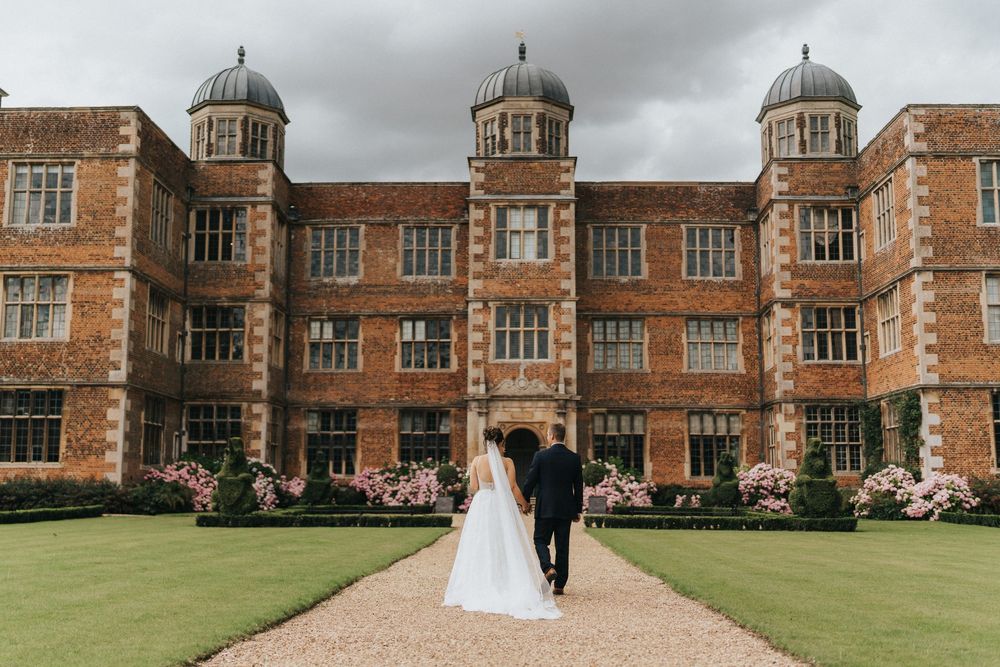 A bride and groom walk along a path beside a grand historic brick manor house with ornate architecture.