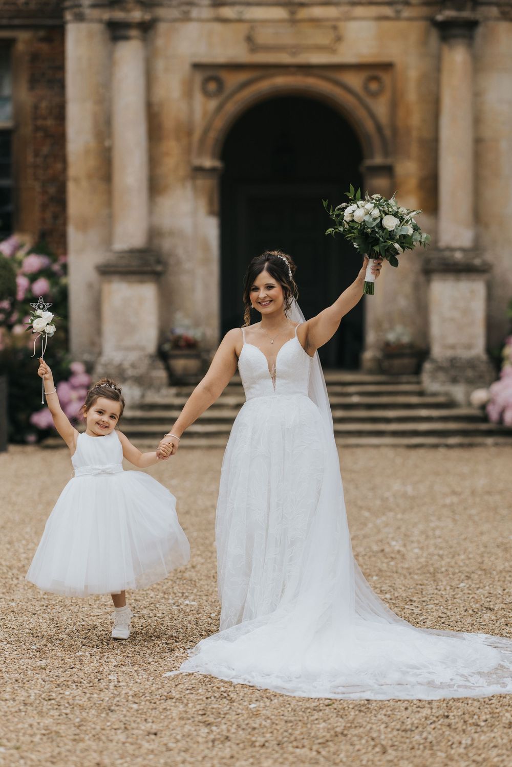 A bride in a flowing white gown holds hands with a young flower girl in a matching white dress outside a grand stone building.