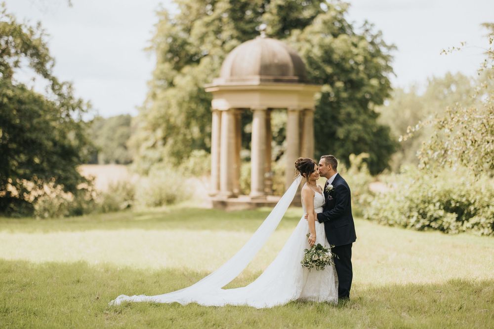 A couple strolls through a lush garden with a classical domed gazebo structure in the background.