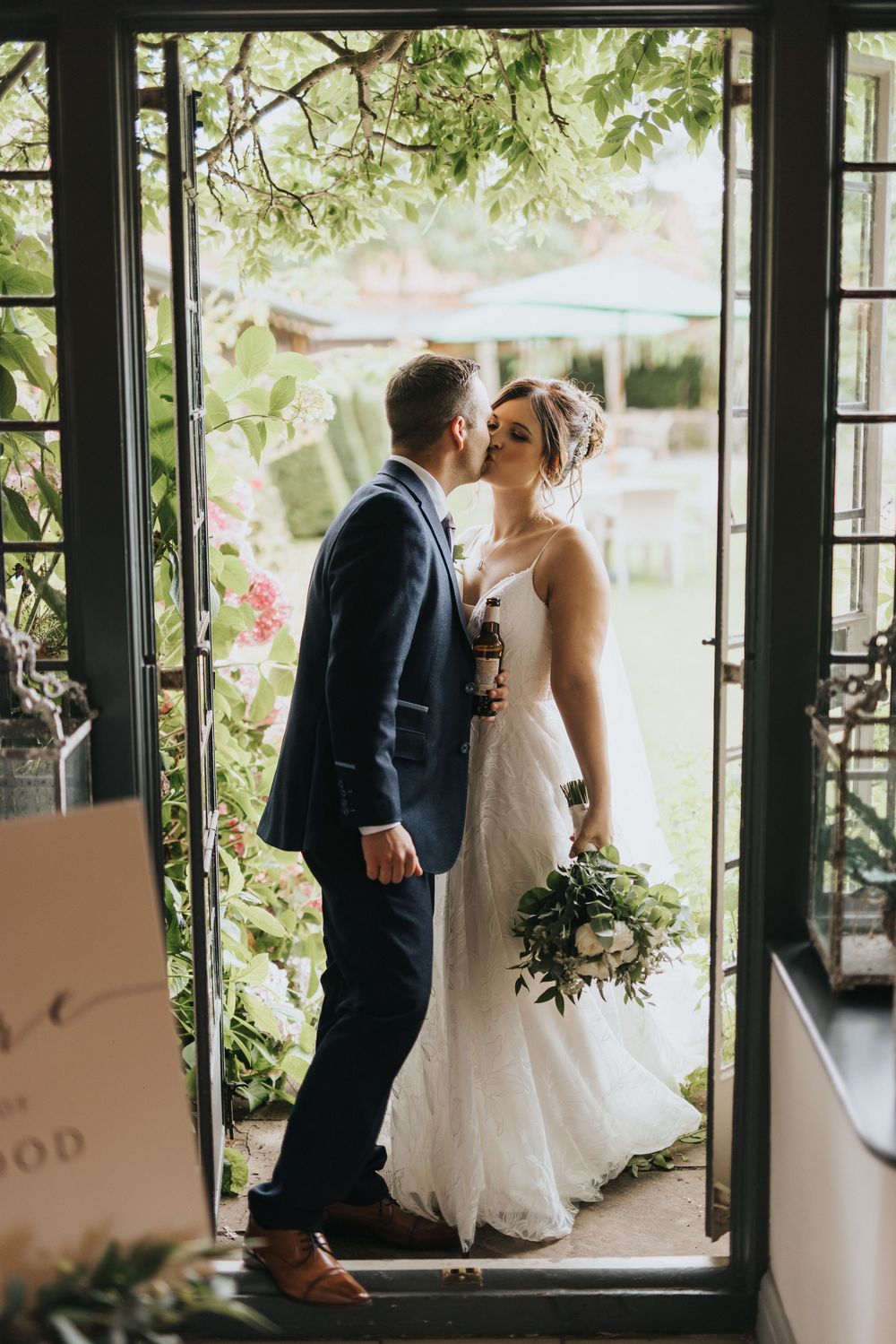 A romantic couple in wedding attire shares a kiss while standing in a doorway framed by greenery and natural light.