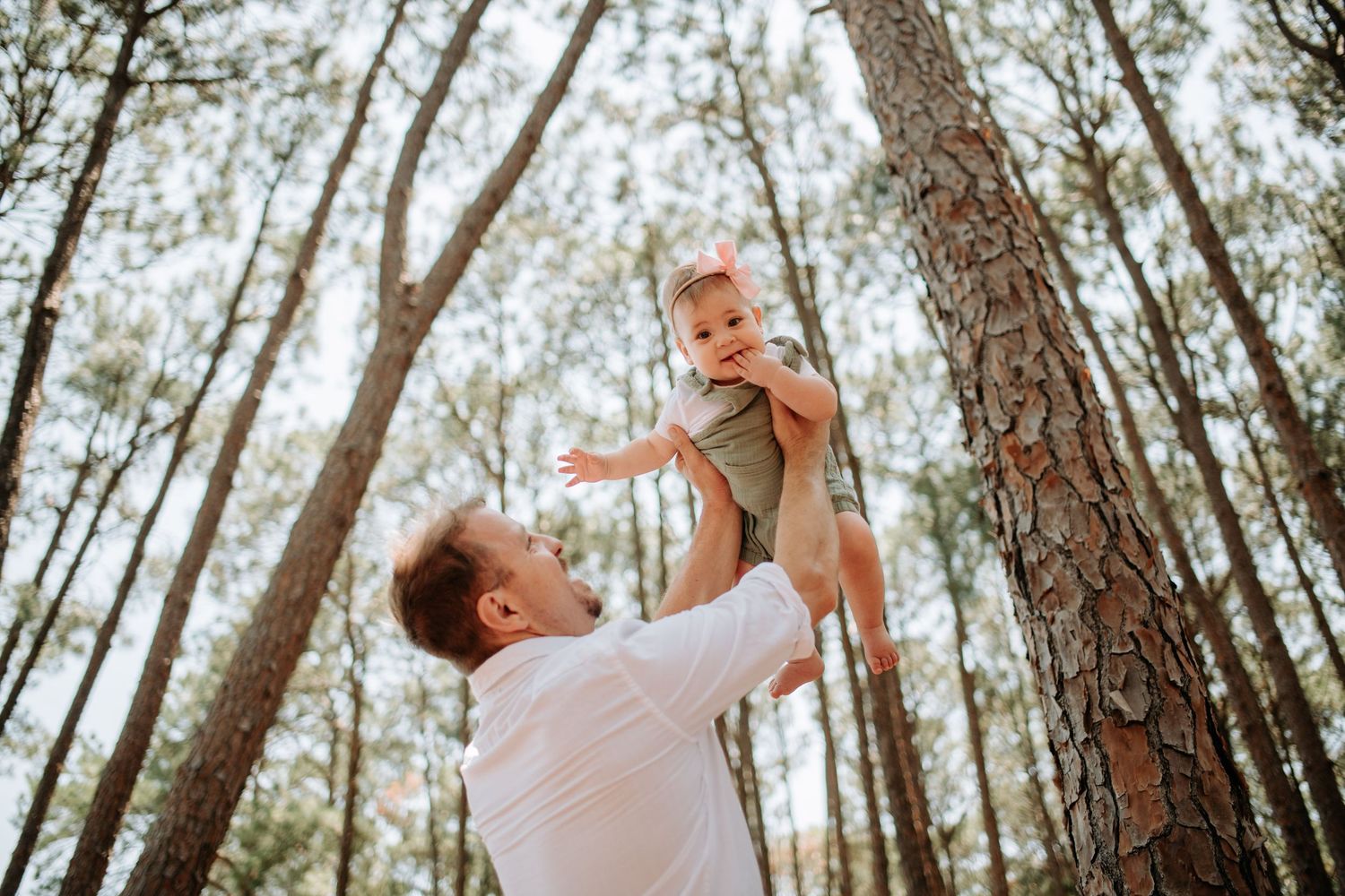 Parent lifts child in the air during joyful moment together among tall pine trees in an outdoor forest setting.