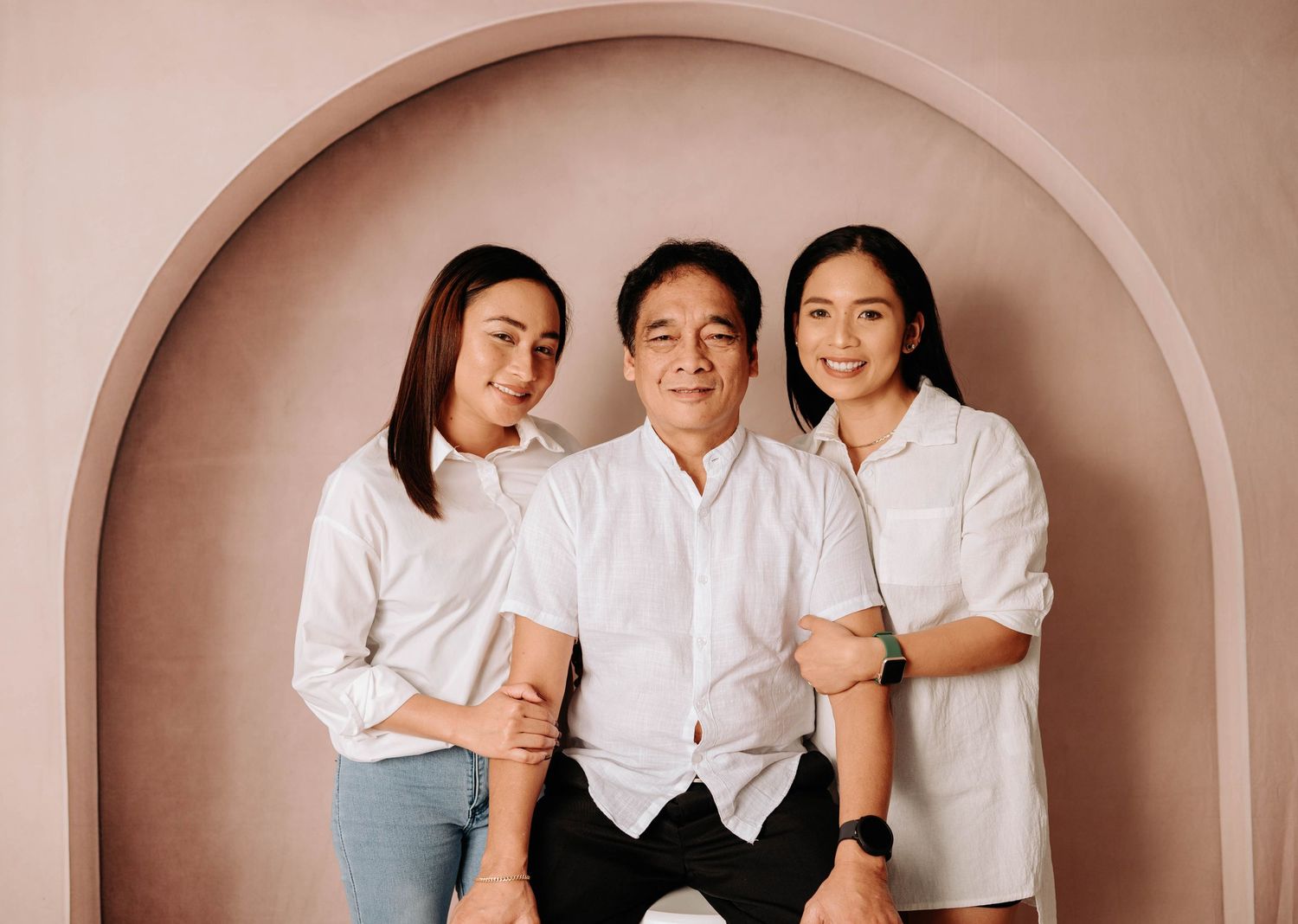 Three people wearing white shirts pose together in front of a beige arched backdrop for a minimalist family portrait.