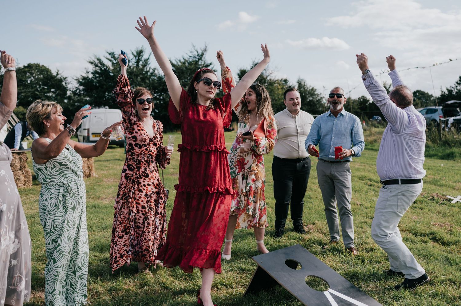 Large group of wedding guests dressed in white and colorful dresses wave their hands in celebration on a grassy field.