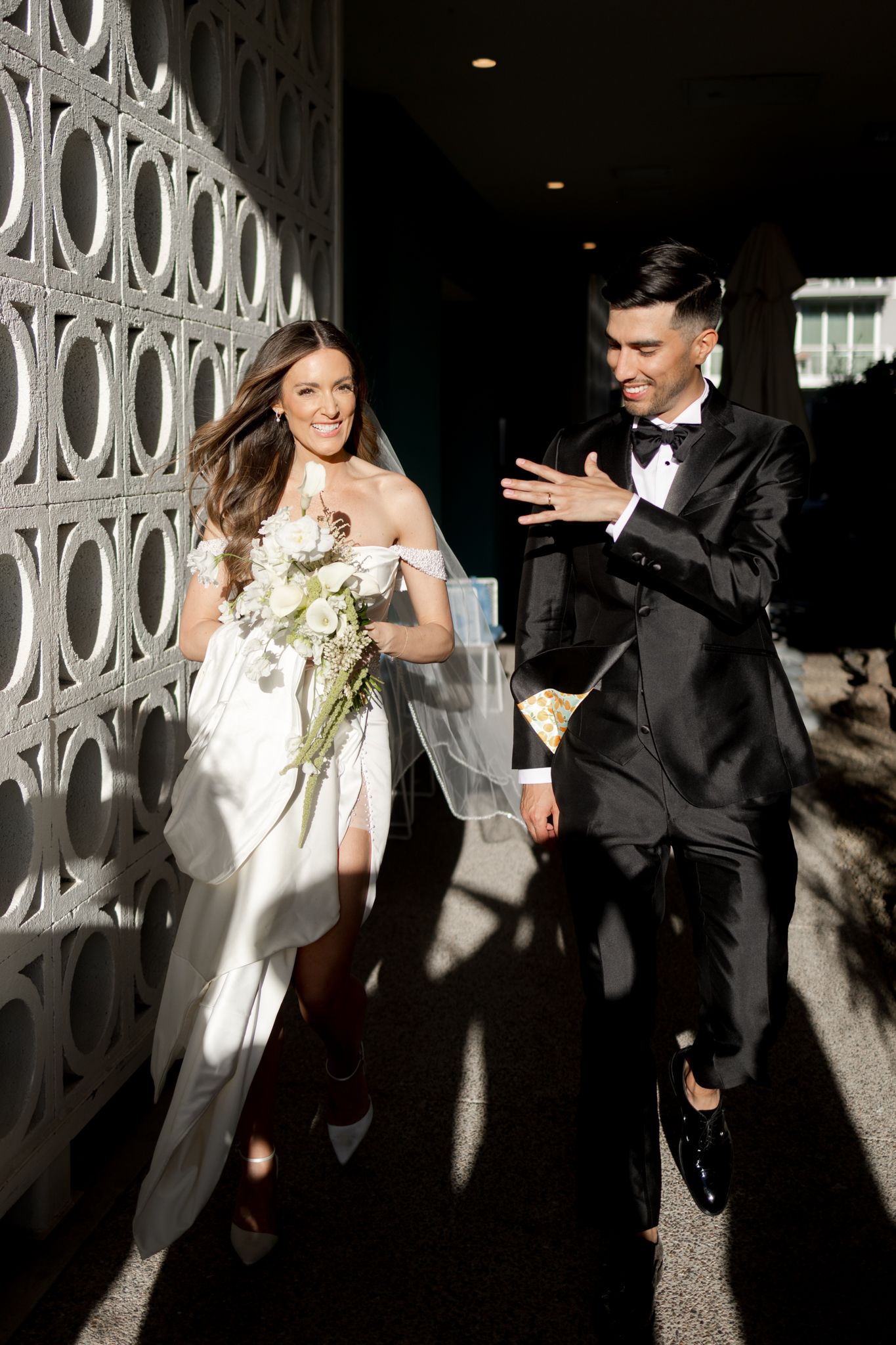 A couple in wedding attire walks and laughs together along a wall with decorative white geometric patterns.