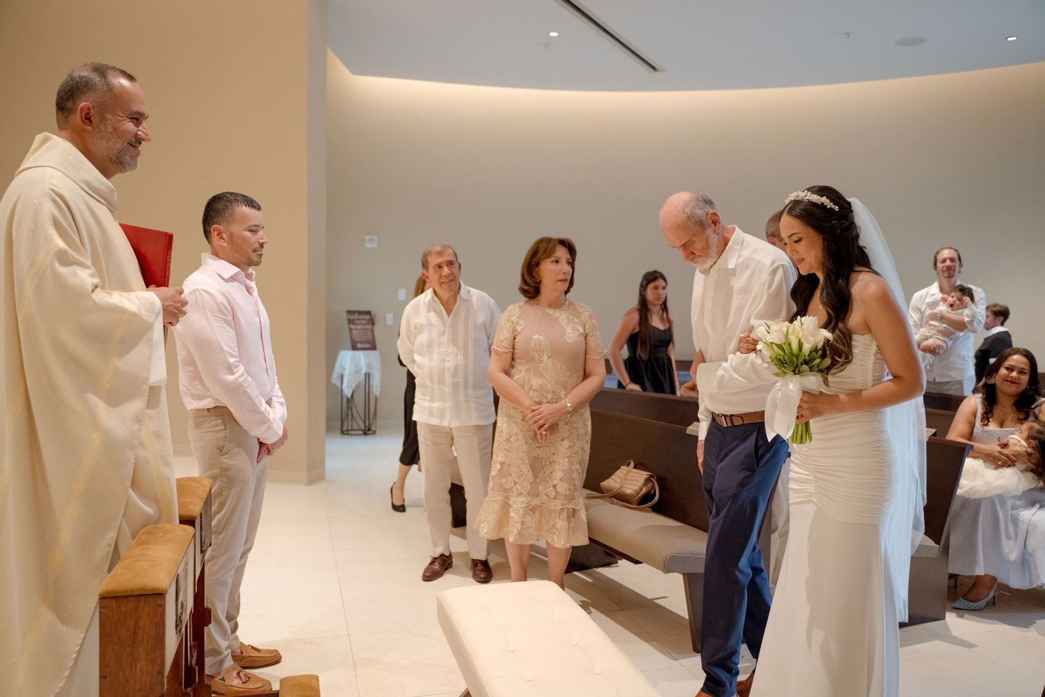 A wedding ceremony in progress inside a modern church with a bride in white dress and guests looking on.