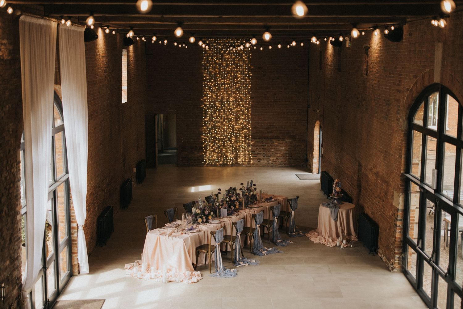 Wedding reception venue with rustic decor features long banquet table and romantic string lights in industrial space.