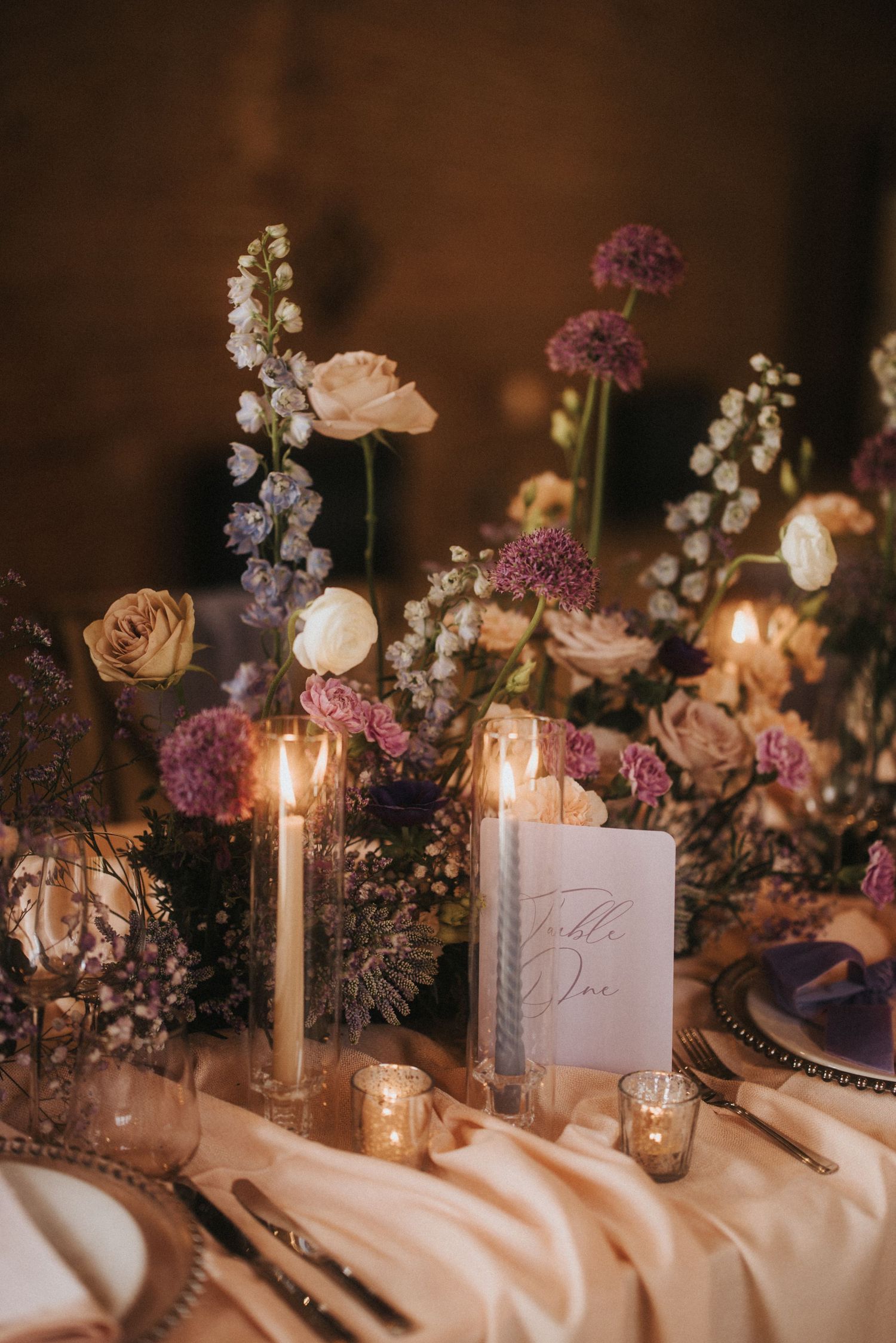Romantic table setting with delicate floral arrangements, lit candles and warm lighting on cream tablecloth.
