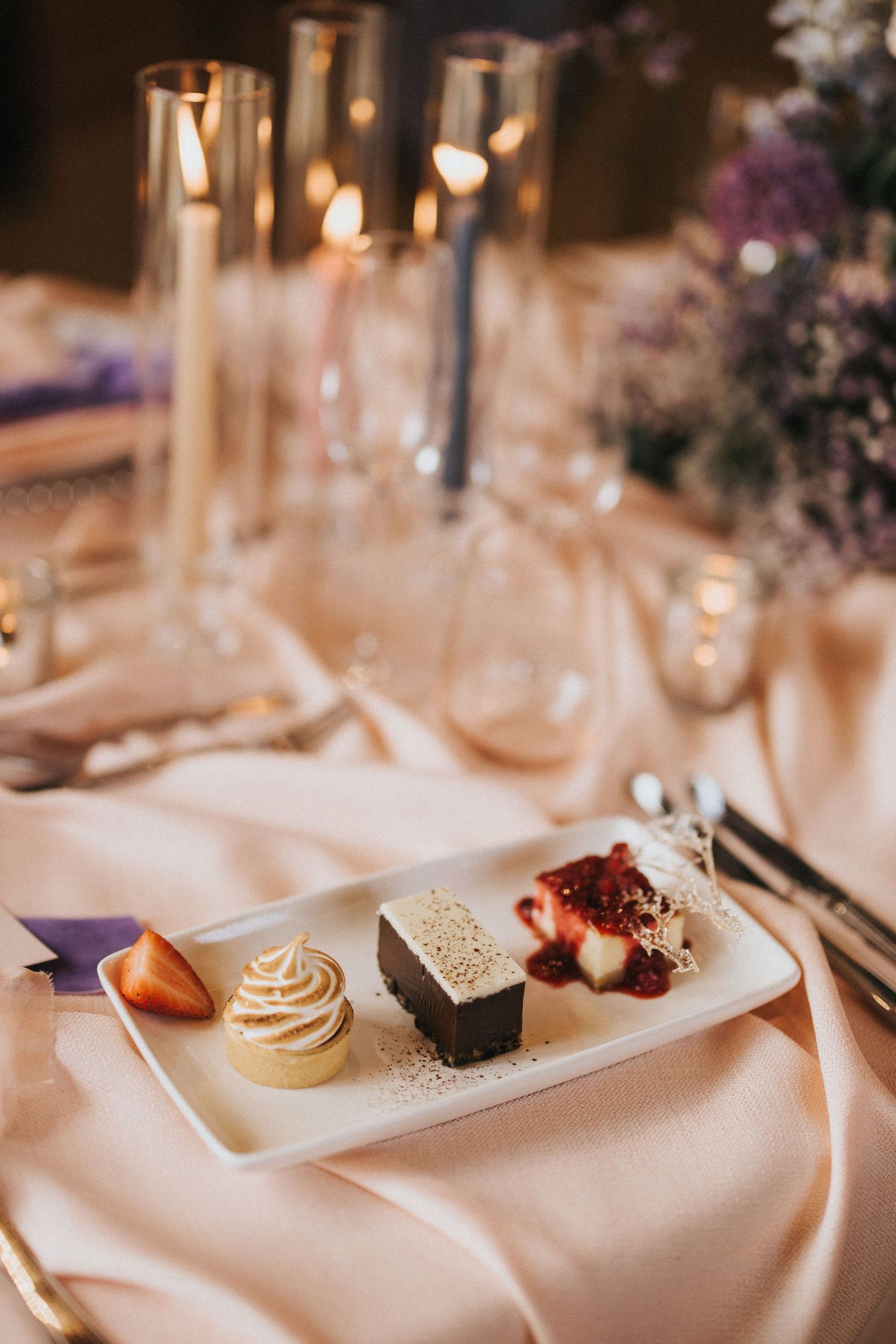 Elegant dessert plate featuring a chocolate brownie, berry sauce, and decorative cupcake on silky tablecloth with glowing candles.