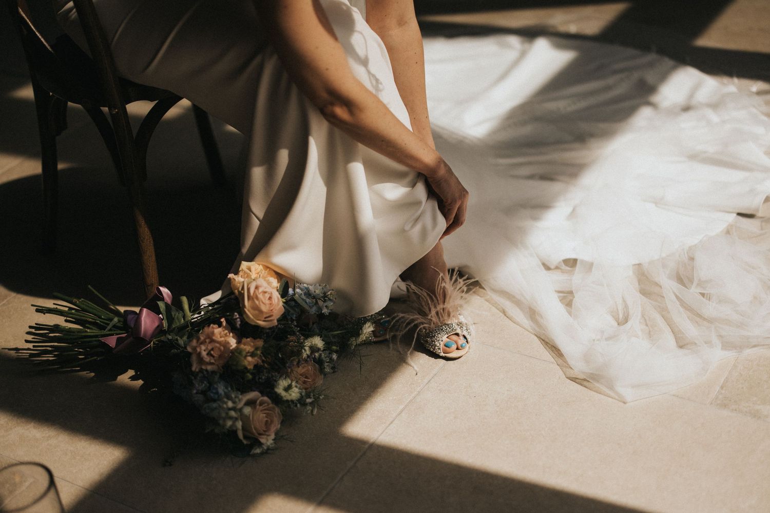 A person in a white dress adjusts their shoes next to a bouquet of flowers on the floor with dramatic shadows.