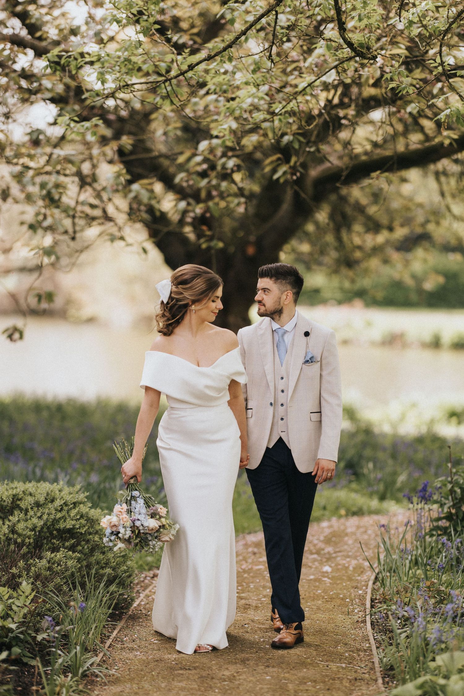 A romantic couple in wedding attire walks together through a beautiful garden path with blooming flowers.