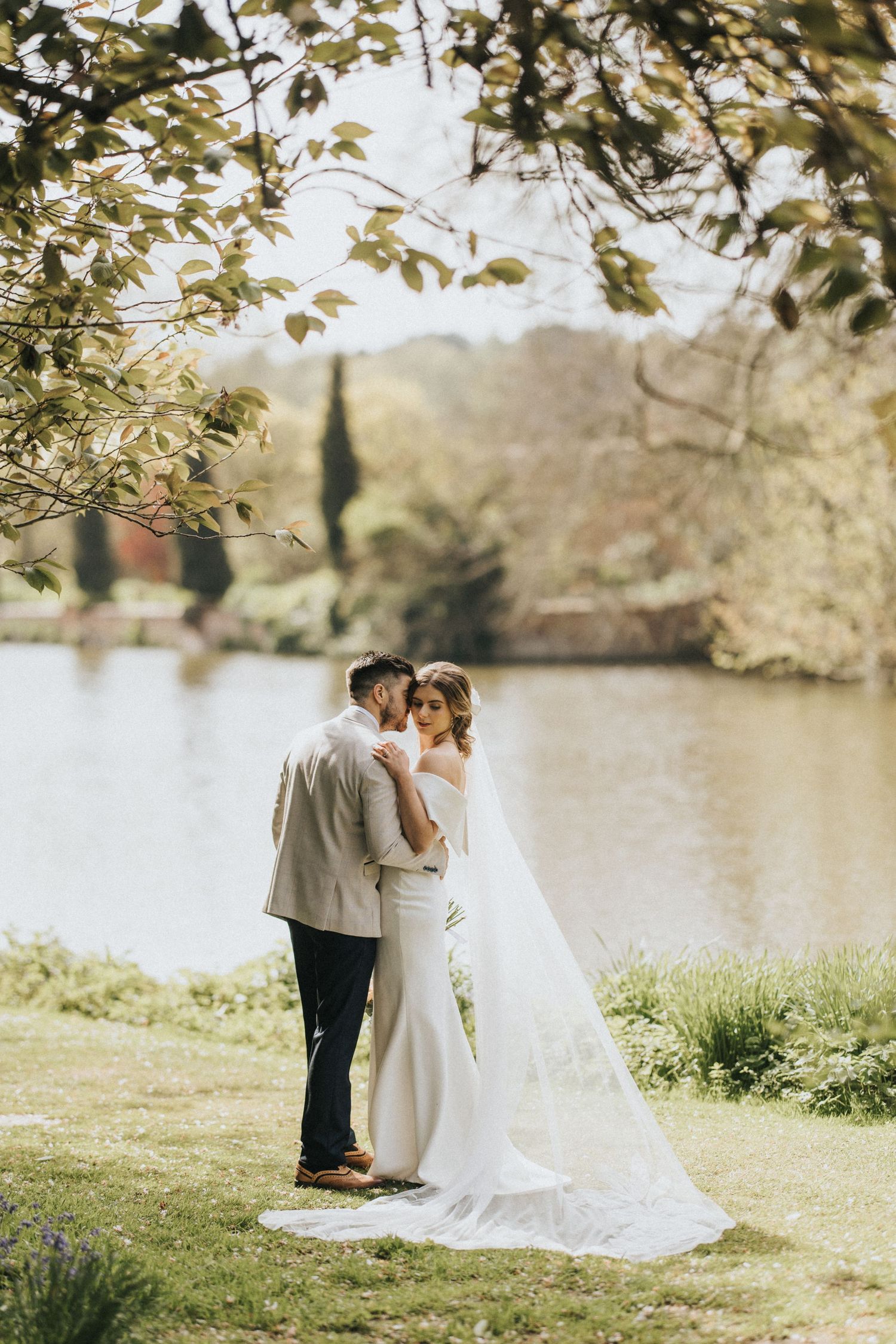 A romantic wedding couple embraces by a serene lakeside on a sunny day, surrounded by green foliage and trees.