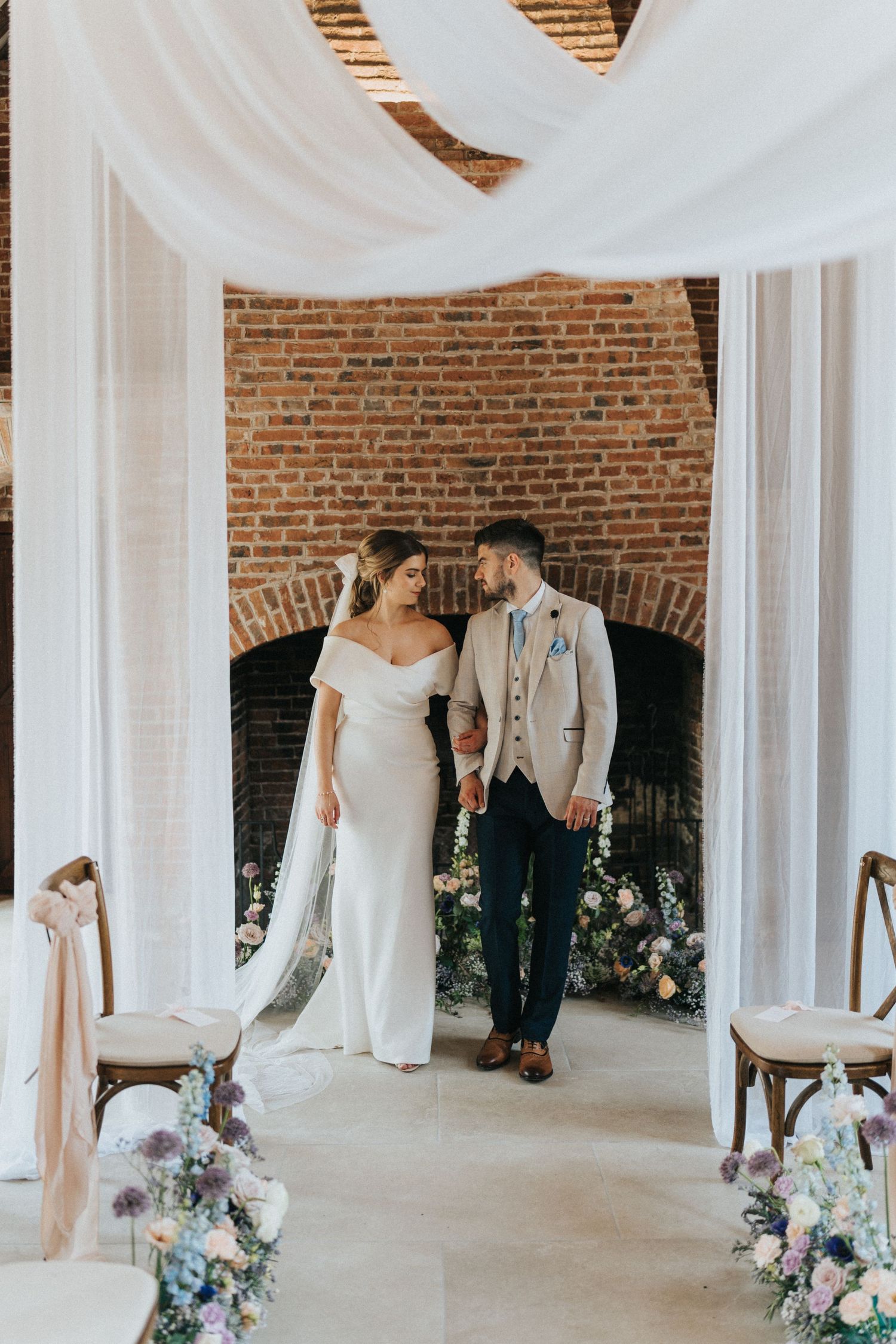 A couple shares an intimate moment under white fabric draping and exposed brick at a rustic indoor wedding venue.