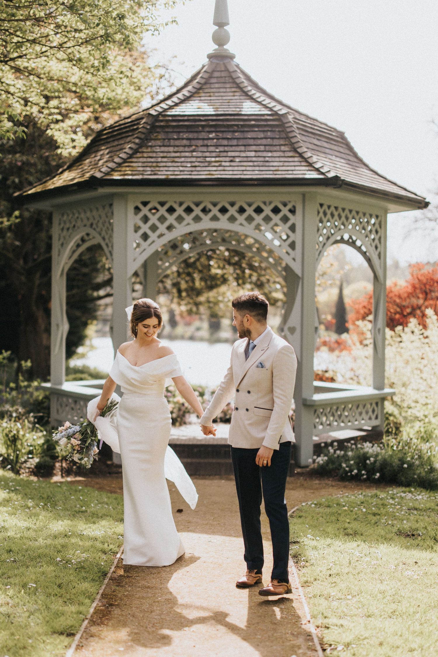 A couple dances playfully under a white garden gazebo on a sunny day surrounded by lush greenery.