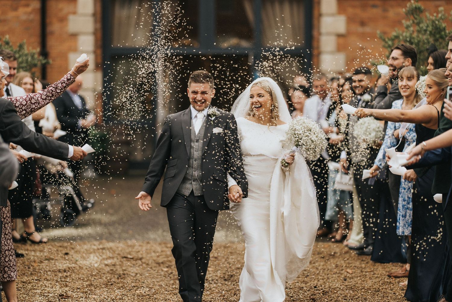 Wedding couple walk through confetti shower from guests at rustic brick venue as they celebrate their special moment.