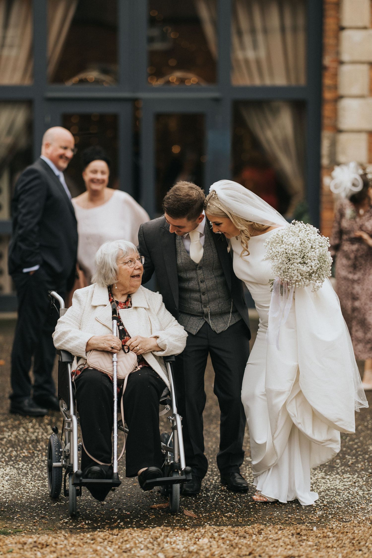 A heartwarming moment at a wedding as a bride in white dress leans down to interact with an elderly guest in a wheelchair.