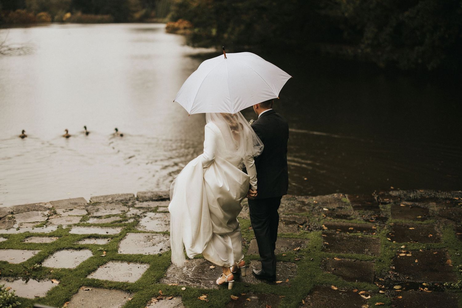 Two people with white umbrella walking together near a lake or pond on an autumn day.