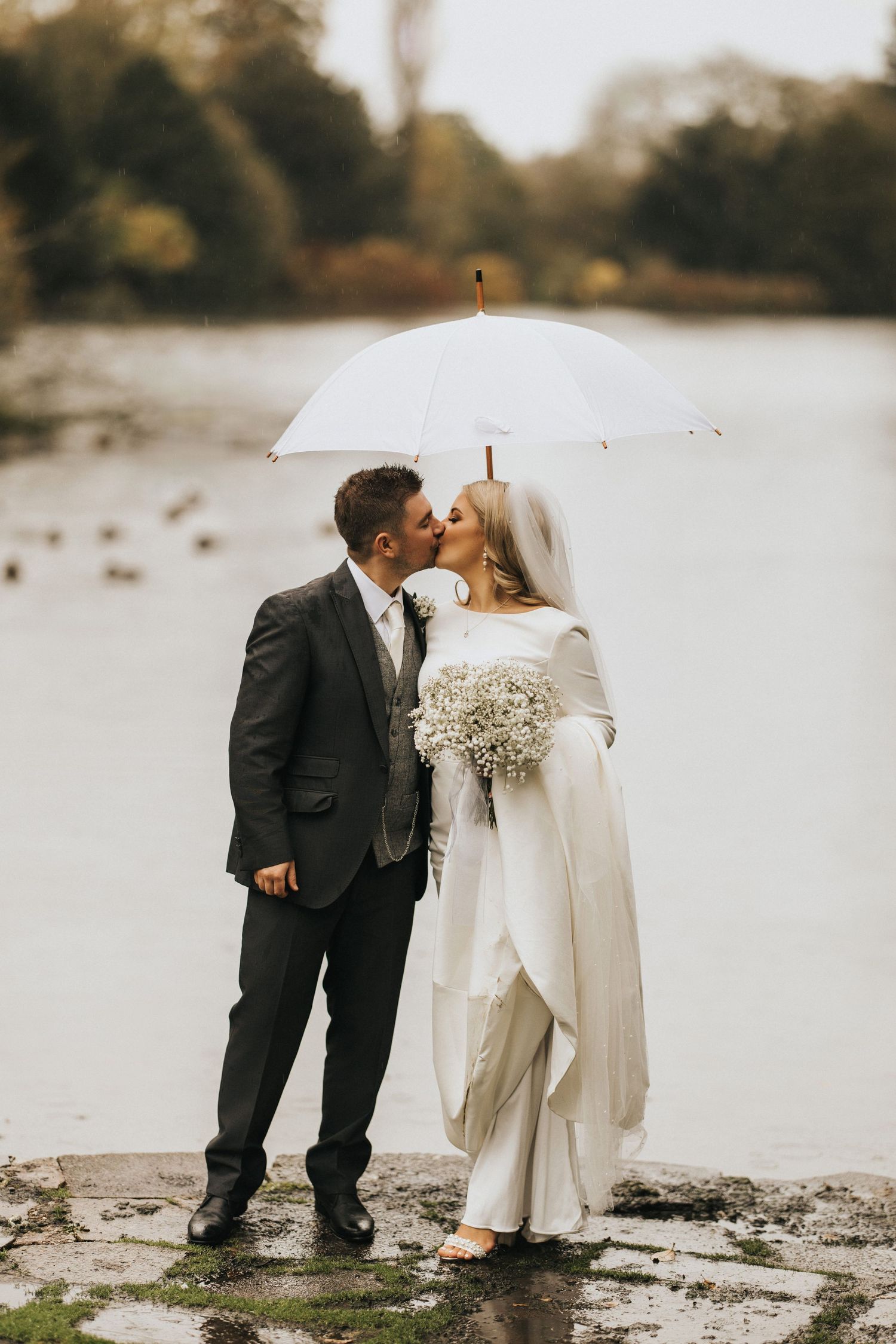 A couple shares a romantic moment under a white umbrella on a rainy day by the lake.