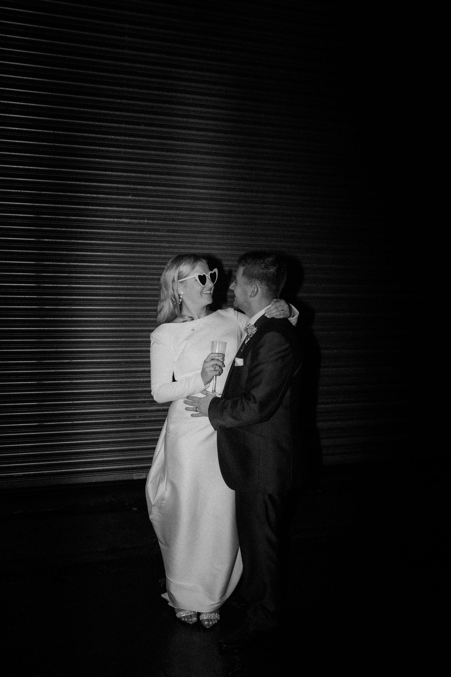 A romantic black and white wedding photo of a couple embracing in front of a striped wall, the bride in white and groom in dark suit.