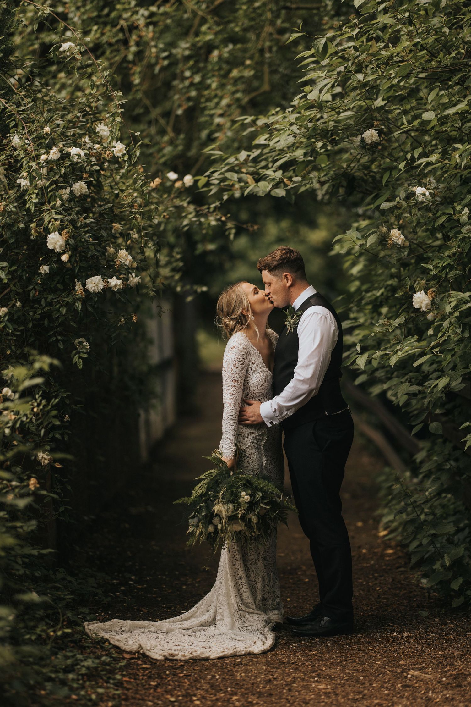 A romantic wedding couple embraces on a garden path surrounded by blooming white roses and lush greenery.