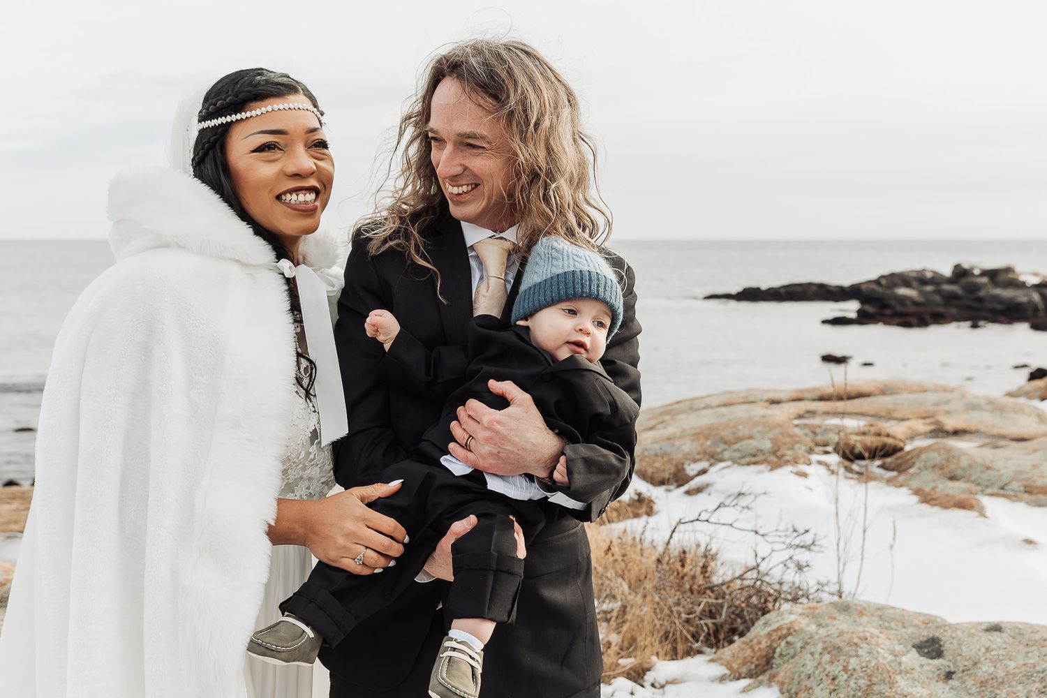 Two people share joyful laughter while holding a baby on a snowy rocky coastline in winter.