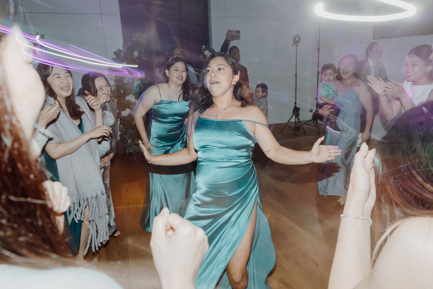 A bridesmaid in a teal satin gown dances energetically on a crowded reception dance floor.