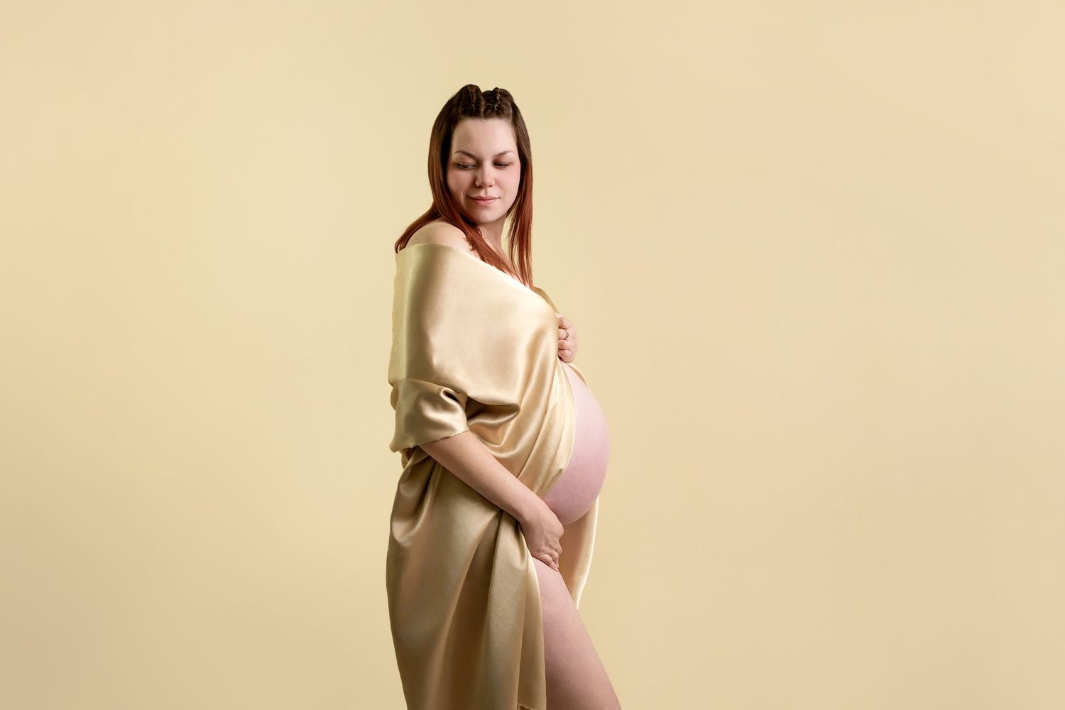 An expectant mother wearing a cream-colored sweater poses against a neutral beige backdrop during a maternity photoshoot.