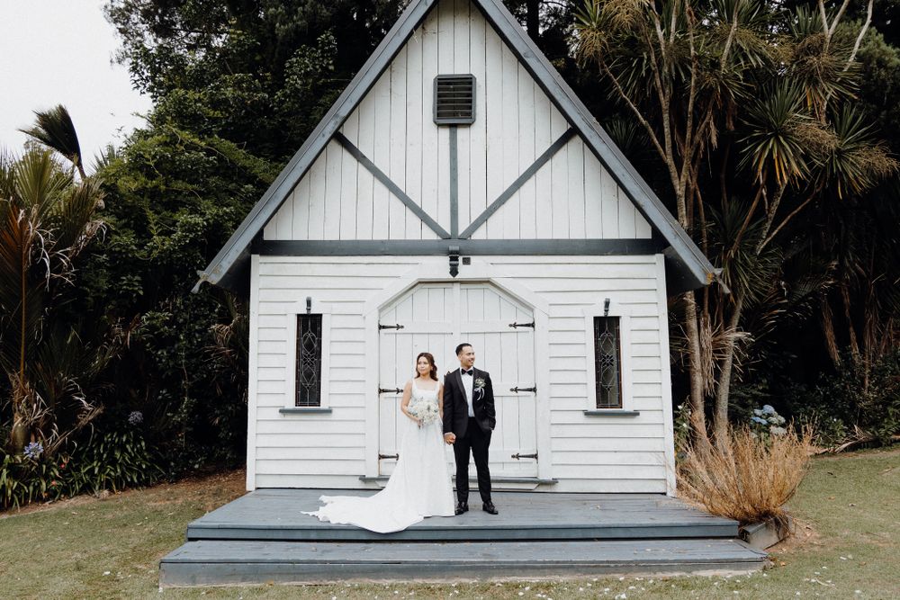 A couple stands together in formal attire in front of a charming white wooden chapel captured by Apertured Media Auckland Wedding Photograph